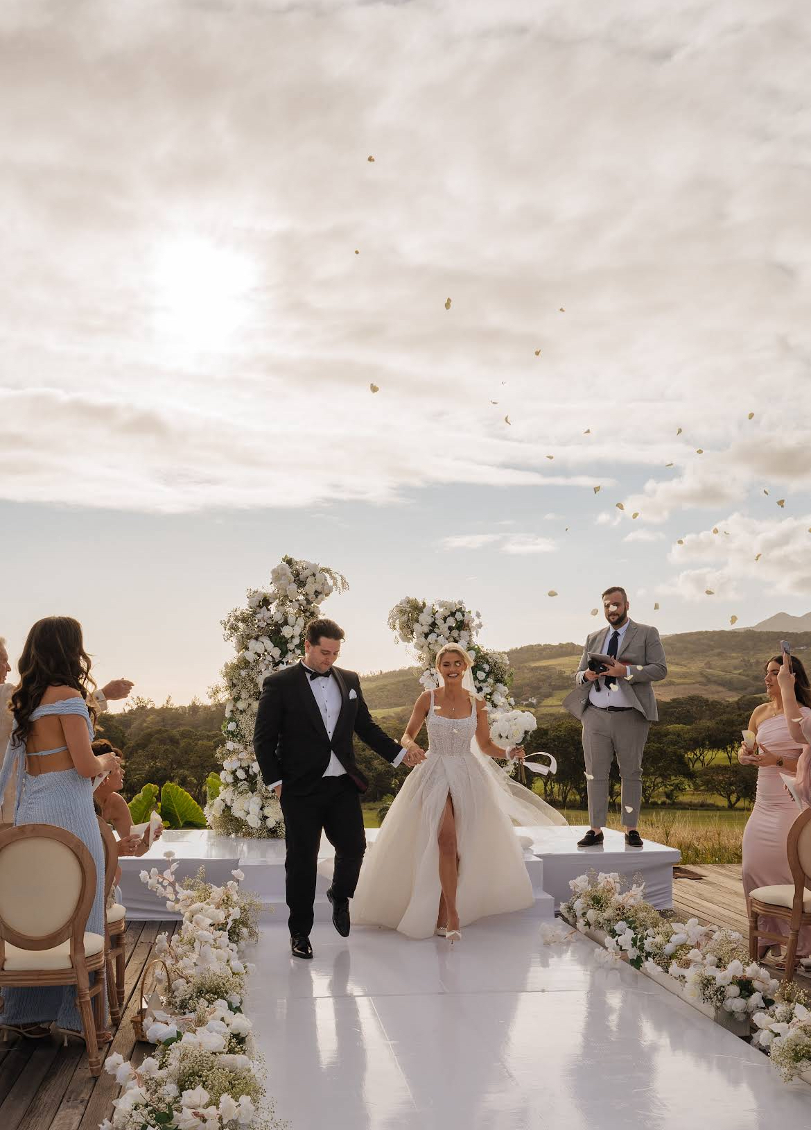 Bride and groom walking down a white aisle lined with white florals at an outdoor wedding ceremony in Mauritius